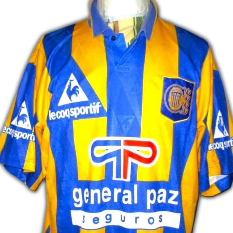 May include: A vintage soccer jersey in blue and yellow, featuring a collared neckline and short sleeves. The jersey has a vertical blue stripe down the center and the text "general paz seguros". The Le Coq Sportif logo is visible.