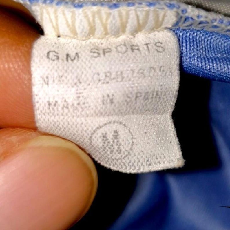 May include: Close-up of a white clothing tag with the text "G.M. SPORTS" and "MADE IN SPAIN". The tag also has the size "M" printed on it. The tag is attached to a blue and gray garment.