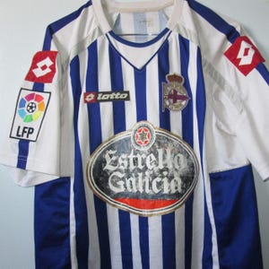 May include: A white and blue striped soccer jersey with the logos of Lotto, LFP, and Estrella Galicia. The jersey has a V-neck and a team emblem on the chest.