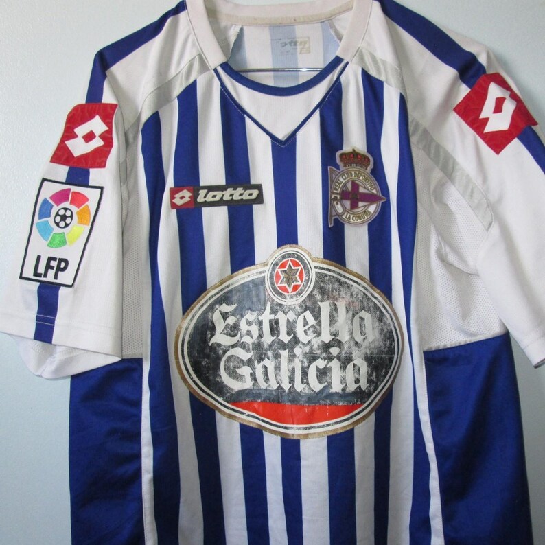 May include: A white and blue striped soccer jersey with the logos of Lotto, LFP, and Estrella Galicia. The jersey has a V-neck and a team emblem on the chest.