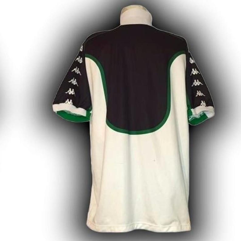 Op de afbeelding: Een wit en zwart shirt met groene rand. Het shirt heeft een zwart bovenpaneel op de rug en groene accenten. De mouwen zijn zwart met witte logo's. De kraag is wit. Dit shirt is waarschijnlijk voor een sportteam.