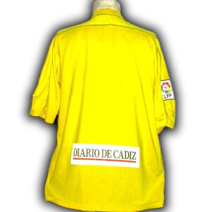 Puede incluir: Camiseta amarilla brillante de manga corta con el texto "DIARIO DE CADIZ" en la espalda. La camiseta tiene un peque&ntilde;o logotipo de la LFP en la manga derecha. La camiseta es de color amarillo liso.