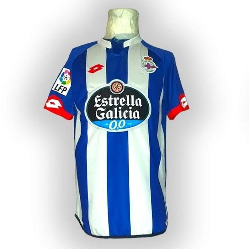 Puede incluir: Una camiseta de f&uacute;tbol a rayas azules y blancas con el logo "Estrella Galicia 0,0" en la parte delantera. La camiseta tiene detalles rojos en las mangas y los logotipos de LFP y Lotto. El escudo del equipo est&aacute; en el hombro derecho.