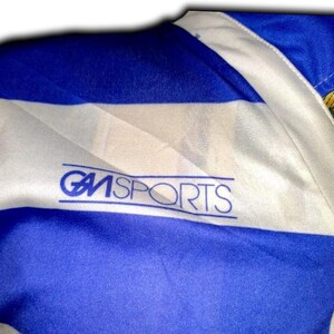 May include: Close-up of a blue and white sports jersey. The jersey has horizontal stripes of blue and white. The text "GM SPORTS" is printed in blue on the white stripe.