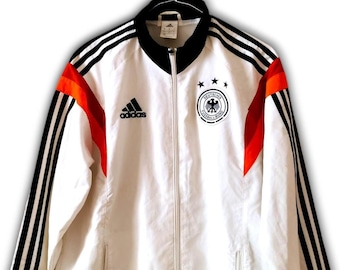 Könnte beinhalten: Weiße Adidas-Trainingsjacke mit schwarzen Streifen auf den Ärmeln und orangefarbenen Akzenten. Mit dem Wappen der deutschen Nationalmannschaft und dem Adidas-Logo. Die Jacke hat einen durchgehenden Reißverschluss und einen schwarzen Kragen.