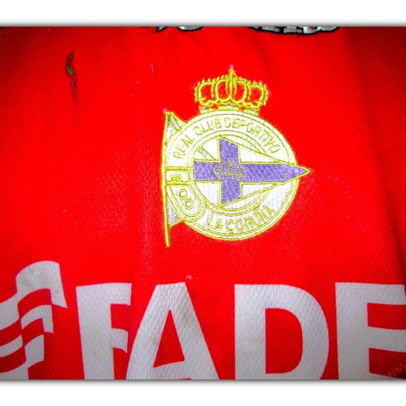 K&ouml;nnte beinhalten: Rotes Trikot mit dem Emblem des Real Club Deportivo La Coru&ntilde;a. Das Emblem ist gold, wei&szlig; und lila und zeigt eine Krone und den Text "REAL CLUB DEPORTIVO LA CORU&Ntilde;A". Das Wort "FADE" ist in Wei&szlig; gedruckt.
