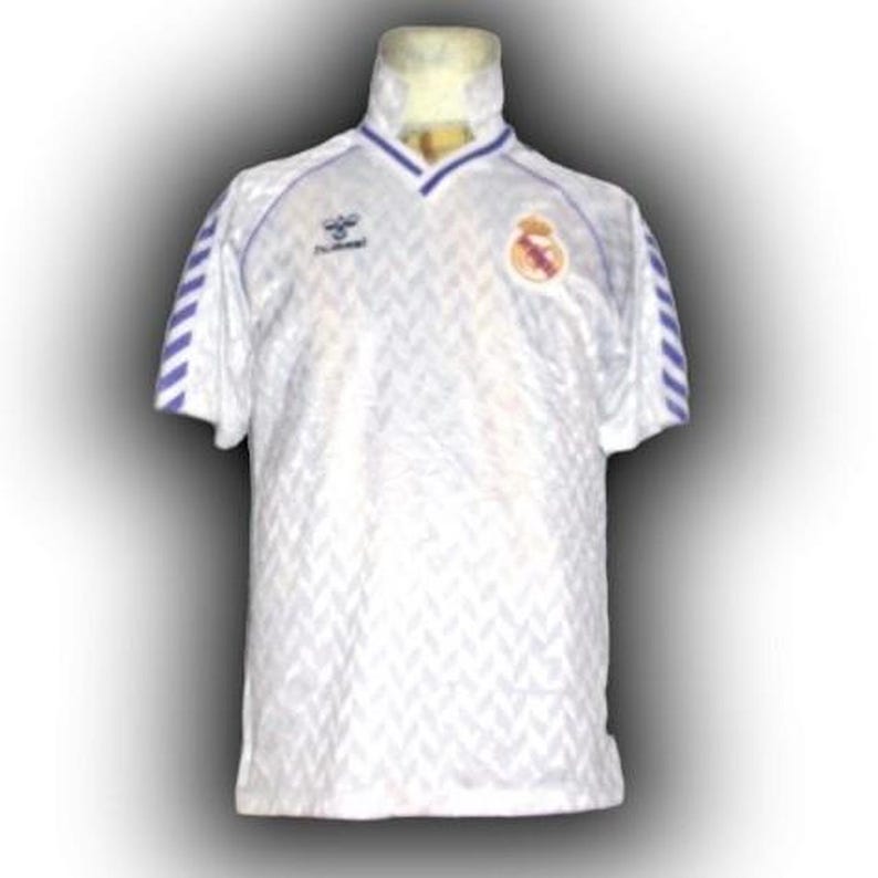 Puede incluir: Camiseta de f&uacute;tbol blanca con un sutil estampado de chevr&oacute;n. Presenta detalles azules en el cuello y las mangas, adem&aacute;s de un escudo del equipo y un logotipo del patrocinador. Manga corta con cuello en V. La camiseta se muestra en un maniqu&iacute;.