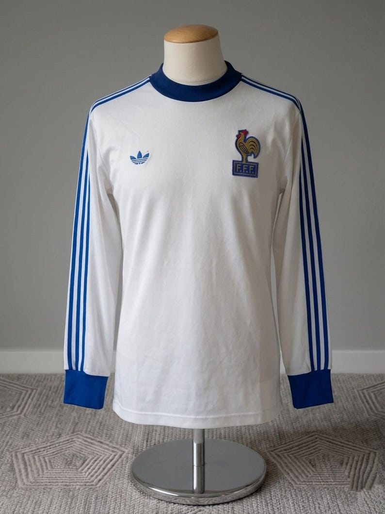 K&ouml;nnte beinhalten: Wei&szlig;es Langarmtrikot mit blauen Akzenten, darunter das Adidas-Logo und das Emblem des franz&ouml;sischen Fu&szlig;ballverbands. Das Trikot hat blaue Streifen auf den &Auml;rmeln sowie einen blauen Kragen und B&uuml;ndchen. Das Trikot wird auf einer Schaufensterpuppe pr&auml;sentiert.
