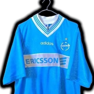 Puede incluir: Una camiseta de f&uacute;tbol Adidas azul claro con cuello y ribetes de manga blancos y azules. La camiseta presenta el logotipo de Adidas, el logotipo de Ericsson y un emblema del equipo. Cuelga de una percha de metal.