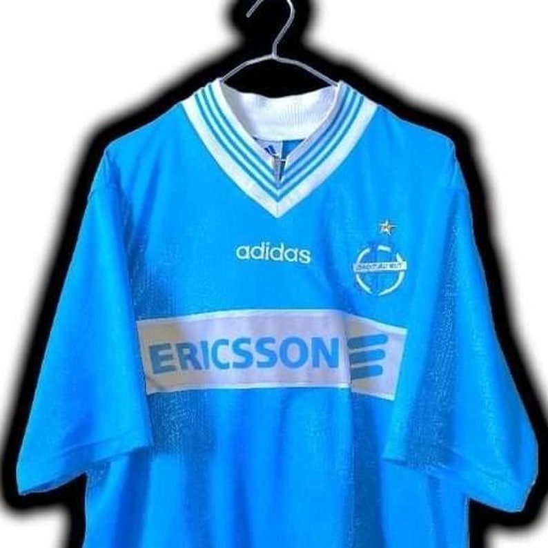 Puede incluir: Una camiseta de f&uacute;tbol Adidas azul claro con cuello y ribetes de manga blancos y azules. La camiseta presenta el logotipo de Adidas, el logotipo de Ericsson y un emblema del equipo. Cuelga de una percha de metal.