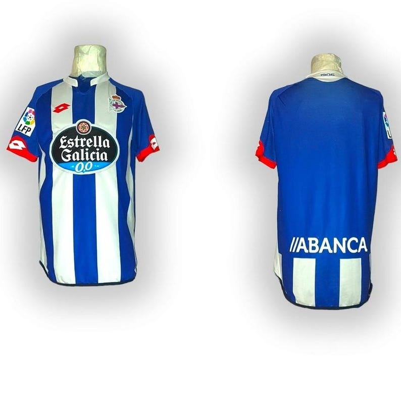 Puede incluir: Una camiseta de f&uacute;tbol a rayas azules y blancas con detalles rojos en las mangas. La parte delantera presenta el logotipo "Estrella Galicia 0.0" y el logotipo de la LFP. La parte trasera de la camiseta tiene la palabra "ABANCA" impresa en blanco.
