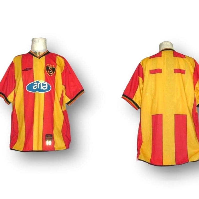 Puede incluir: Una camiseta de f&uacute;tbol vintage, con un dise&ntilde;o a rayas rojas y amarillas con ribete negro. La camiseta tiene cuello en V y la palabra "aria" en un &oacute;valo azul. La parte trasera de la camiseta tambi&eacute;n es visible.
