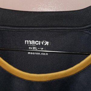 Puede incluir: Camiseta deportiva negra con un ribete dorado alrededor del cuello. El logotipo de Macron es visible en blanco, junto con el texto "EU XL-IR" y "macron.com". La camiseta tiene un cuello acanalado y parece estar hecha de una tela transpirable.