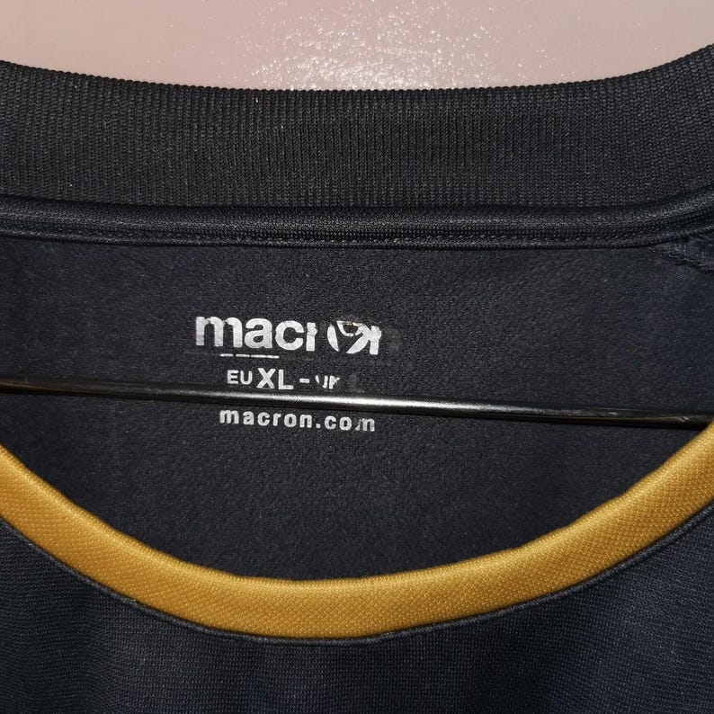 Puede incluir: Camiseta deportiva negra con un ribete dorado alrededor del cuello. El logotipo de Macron es visible en blanco, junto con el texto "EU XL-IR" y "macron.com". La camiseta tiene un cuello acanalado y parece estar hecha de una tela transpirable.