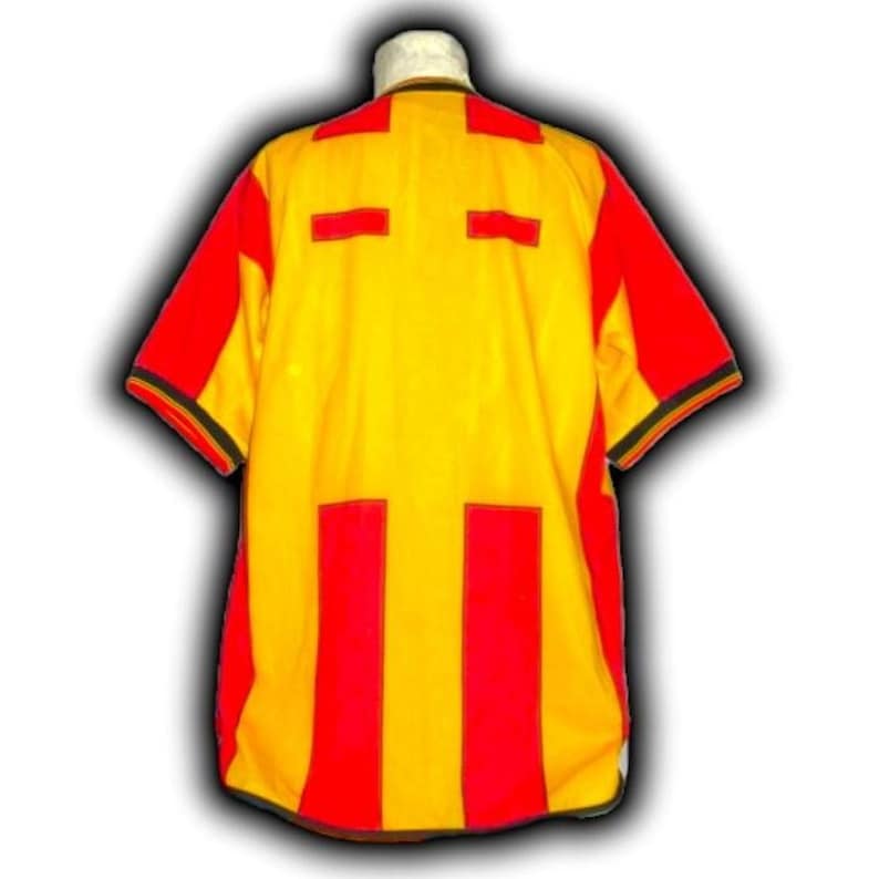Puede incluir: Una camiseta amarilla y roja de manga corta. La camiseta tiene paneles rojos en los hombros y las mangas, con rayas verticales rojas y detalles rectangulares en la parte delantera y trasera. El cuello es amarillo con ribete negro.