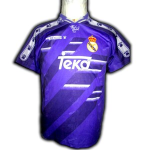 K&ouml;nnte beinhalten: Violett-wei&szlig;es Fu&szlig;balltrikot mit dem Wort "Teka" auf der Vorderseite. Das Trikot hat einen Kragen und kurze &Auml;rmel. Das Teamlogo befindet sich auf der Brust, und Pfotenabdr&uuml;cke verzieren die Schultern.