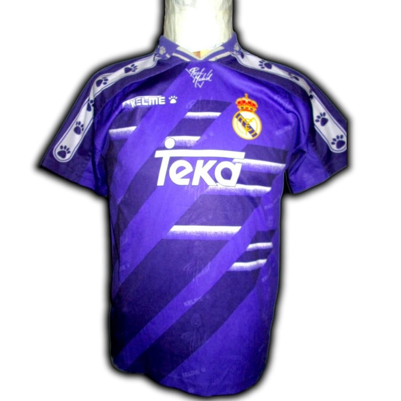 K&ouml;nnte beinhalten: Violett-wei&szlig;es Fu&szlig;balltrikot mit dem Wort "Teka" auf der Vorderseite. Das Trikot hat einen Kragen und kurze &Auml;rmel. Das Teamlogo befindet sich auf der Brust, und Pfotenabdr&uuml;cke verzieren die Schultern.