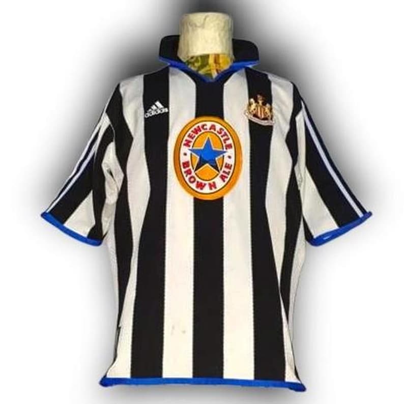 Op de afbeelding: Zwart-wit gestreept voetbalshirt met blauwe accenten. Voorzien van het Adidas-logo, een cirkelvormig embleem met een blauwe ster en de tekst "Newcastle Brown Ale", en een teamembleem.