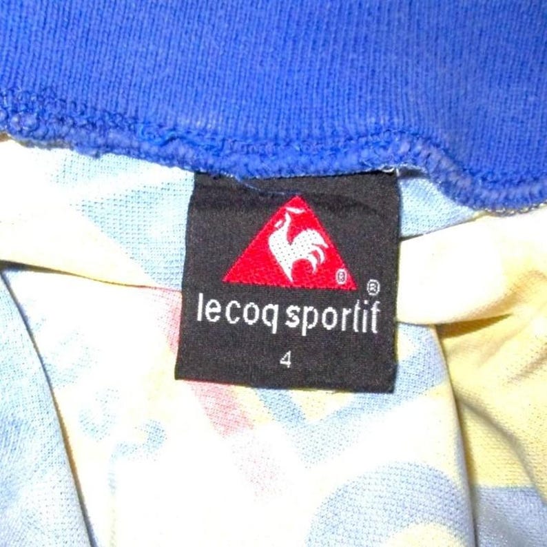May include: Close-up of a black label with the text "le coq sportif" in white, and a red triangle logo with a white rooster. The label is on a patterned fabric with blue and yellow hues, and a blue collar is visible.