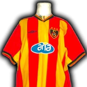 Puede incluir: Una camiseta de f&uacute;tbol a rayas rojas, amarillas y negras con la palabra "aria" en un &oacute;valo azul en la parte delantera. La camiseta tiene cuello en V y un logotipo del equipo en el pecho.