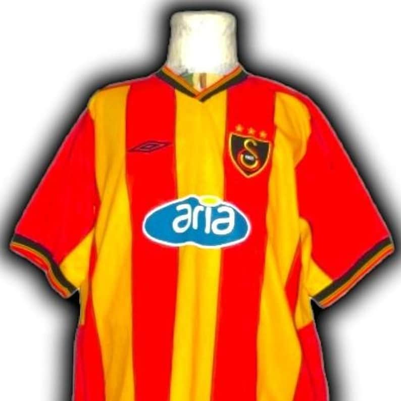 Puede incluir: Una camiseta de f&uacute;tbol a rayas rojas, amarillas y negras con la palabra "aria" en un &oacute;valo azul en la parte delantera. La camiseta tiene cuello en V y un logotipo del equipo en el pecho.