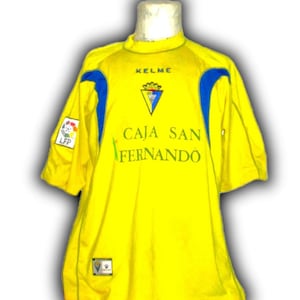 Puede incluir: Una camiseta de f&uacute;tbol amarilla con detalles azules en los hombros. La camiseta presenta el logo "KELME", el texto "CAJA SAN FERNANDO" y el logo LFP. Tiene mangas cortas y cuello redondo.