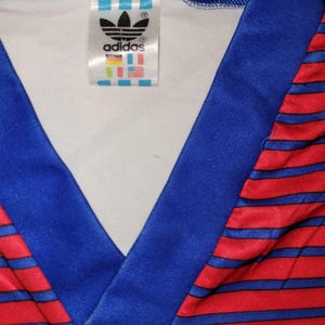May include: Close-up of a vintage Adidas jersey. The jersey features a blue V-neck collar and red and blue striped pattern. The Adidas logo and brand tag are visible on the inside of the collar.