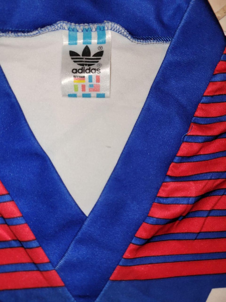 May include: Close-up of a vintage Adidas jersey. The jersey features a blue V-neck collar and red and blue striped pattern. The Adidas logo and brand tag are visible on the inside of the collar.