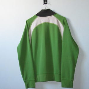 K&ouml;nnte beinhalten: Gr&uuml;ne Jacke mit Rei&szlig;verschluss, schwarzem Kragen und wei&szlig;en Schulterpartien. Die Jacke h&auml;ngt an einem schwarzen Haken vor einer wei&szlig;en Wand. Die &Auml;rmel sind lang und die B&uuml;ndchen gerippt.