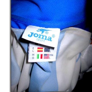 May include: Close-up of a blue and white athletic jersey with the "Joma sport" logo on a tag. The tag also displays flags of different countries and size labels.
