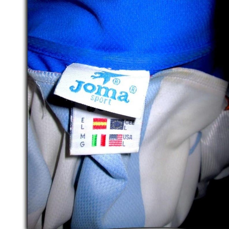 May include: Close-up of a blue and white athletic jersey with the "Joma sport" logo on a tag. The tag also displays flags of different countries and size labels.