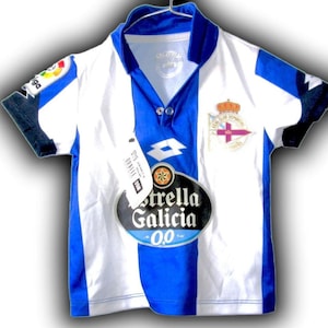 May include: White and blue soccer jersey with a blue vertical stripe down the center. The jersey has a collar and short sleeves with black trim. Logos include "Estrella Galicia 0.0", "Lotto", and a team crest.