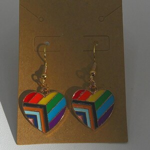 May include: A pair of gold-toned hook earrings featuring heart-shaped pendants. Each heart is decorated with a geometric pattern of enamel stripes in red, yellow, green, blue, purple, orange, and light blue, attached to gold-toned metal hooks.