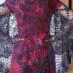 Pode incluir: Um xale de crochê azul marinho com um delicado design vazado. O xale é drapeado sobre uma peça de roupa vermelha, criando um contraste de cores e texturas. O padrão intrincado e a forma fluida sugerem uma peça artesanal, feita à mão, adequada para sobreposição.
