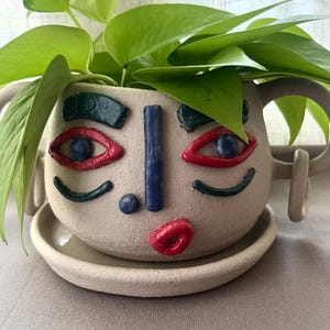 Könnte beinhalten: Ein beigefarbener Keramik-Pflanztopf mit einem Gesichtsmotiv, bestehend aus dunkelgrünen Augenbrauen, rot umrandeten Augen mit blauen Pupillen, einer blauen vertikalen Nase, einem kleinen blauen Punkt und roten Lippen. Er hat zwei seitliche Henkel.