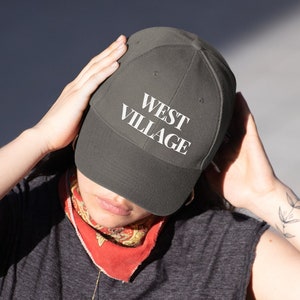 May include: A person wearing a dark gray t-shirt and black pants with a green baseball cap that says "WEST VILLAGE" in white lettering.