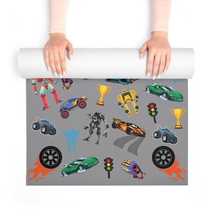 Puede incluir: Una esterilla de yoga gris con ilustraciones coloridas de coches de carreras, robots, trofeos y semáforos. La esterilla tiene un patrón repetido de varios vehículos y gráficos temáticos de carreras.
