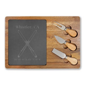 Op de afbeelding: Een houten kaasplank met een donkergrijze leisteen inzet. De leisteen is geëtst met gekruiste ski's, de tekst "Whistler, CA" en coördinaten. Drie kaasmesjes met houten handvatten zijn inbegrepen.