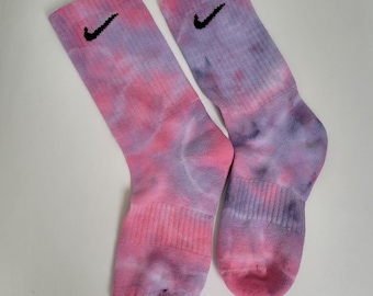 Puede incluir: Un par de calcetines tie-dye rosa y morado con un logotipo Nike negro en forma de swoosh en cada calcetín.