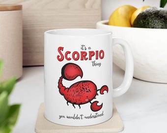 May include: White ceramic mug featuring a red scorpion illustration with the text "it's a Scorpio thing, you wouldn't understand." The mug has a curved handle and is sitting on a beige coaster.