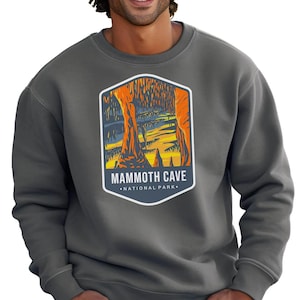 A person wears a Mammoth Cave National Park sweatshirt. The design shows vibrant cave formations in orange and yellow, reflecting the park's caves. The person smiles with hands resting on their knees.