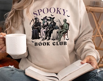 Puede incluir: Una sudadera beige con un gráfico de un grupo de personajes espeluznantes leyendo en un club de lectura. El texto "Spooky Book Club" está impreso en la sudadera.