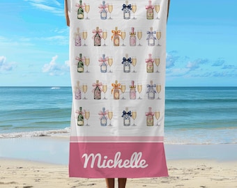 May include: A white beach towel with a pink border and the name "Michelle" printed on the bottom. The towel features a repeating pattern of champagne bottles and glasses.