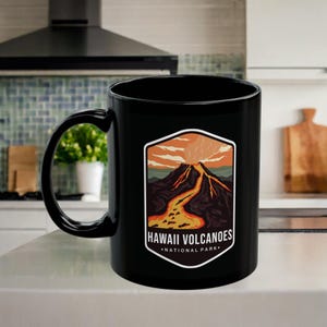 Könnte beinhalten: Schwarze Keramik-Kaffeetasse mit einer Grafik eines Vulkans, der ausbricht und Lava an den Seiten herunterfließt. Der Text "Hawaii Volcanoes National Park" ist auf die Tasse gedruckt.