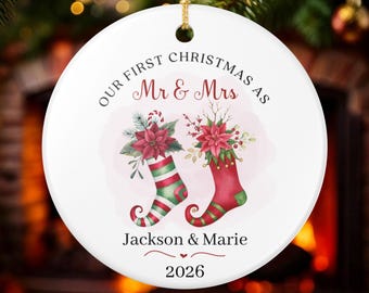 Puede incluir: Un adorno redondo de cerámica blanca con el texto "OUR FIRST CHRISTMAS AS Mr & Mrs" sobre una ilustración en acuarela de dos calcetines navideños llenos de flores de pascua. Debajo aparecen los nombres "Jackson & Marie" y el año "2026".