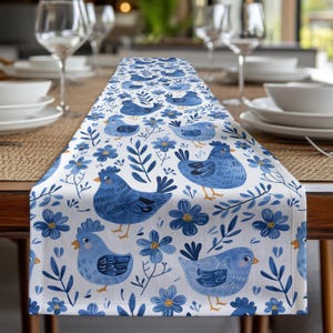 May include: A white table runner with a blue and white floral pattern featuring blue chickens and leaves. The runner is on a wooden table with place settings.