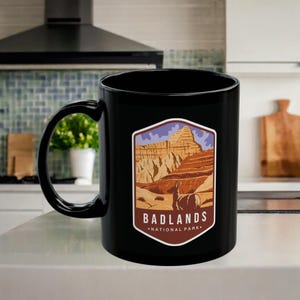 May include: Black ceramic coffee mug with a graphic of Badlands National Park, featuring a mountain range and a bison. The text "Badlands National Park" is printed below the graphic.