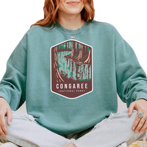 May include: A teal blue sweatshirt with a brown graphic of a river flowing through a forest. The graphic has the text "Congaree National Park" underneath.