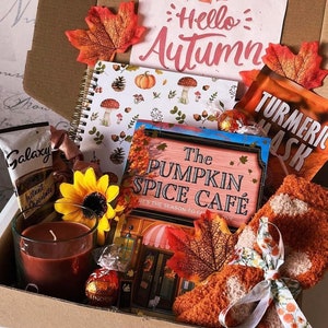 May include: An autumn-themed gift box containing a variety of items. Inside are a notebook with mushroom and pumpkin designs, a candle, a face mask, socks, and a card reading "Hello Autumn". Also included are a chocolate bar and a book titled "The Pumpkin Spice Cafe".