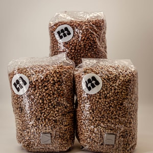 Könnte beinhalten: Drei transparente Plastiktüten gefüllt mit braunen Körnern, pyramidenförmig gestapelt. Jede Tüte hat einen weißen runden Aufkleber mit einem schwarzen Logo. Die Tüten sind versiegelt und haben unten ein kleines graues Quadrat.